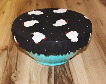 May include: A black fabric bowl cover with a white polka dot pattern and a white chicken design. The cover is sitting on top of a turquoise bowl.