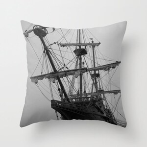 May include: A black and white photograph of a tall ship with multiple masts and sails. The ship is docked and the image is taken from a low angle, looking up at the ship.