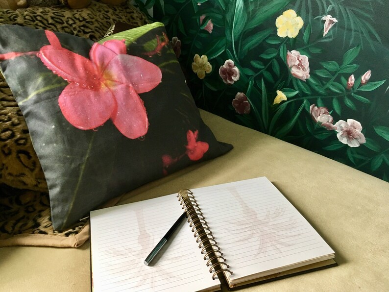 Puede incluir: Una almohada floral en blanco y negro con una flor rosa en un sof&aacute; con un cuaderno y un bol&iacute;grafo encima. El sof&aacute; est&aacute; frente a una pared verde con un patr&oacute;n floral.