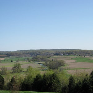 May include: A panoramic view of a rural landscape with rolling hills, green fields, and a winding river. The sky is a clear blue, and the trees are a mix of green and brown.