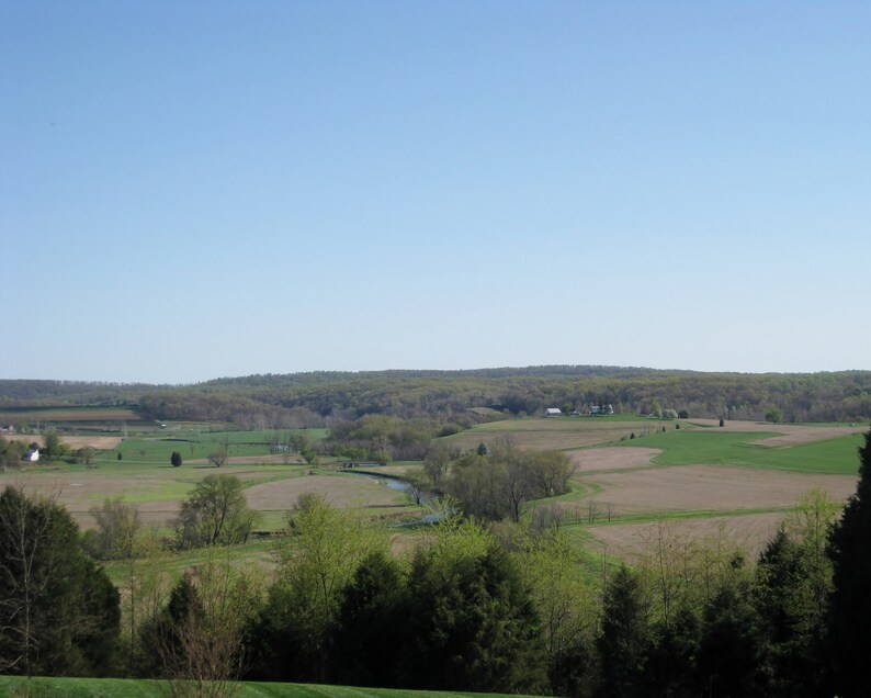 May include: A panoramic view of a rural landscape with rolling hills, green fields, and a winding river. The sky is a clear blue, and the trees are a mix of green and brown.