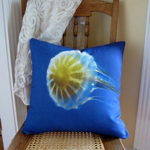 May include: A blue pillow with a white jellyfish with a yellow center on it. The pillow is sitting on a wooden chair with a woven seat.