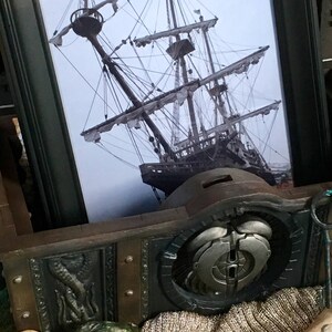 Puede incluir: Una fotograf&iacute;a en blanco y negro enmarcada de un barco alto con velas, sobre un fondo de madera con una pieza de metal decorativa y una botella de l&iacute;quido verde con un tap&oacute;n de corcho.