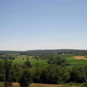 May include: A panoramic view of a rural landscape with rolling green hills, fields, and trees under a clear blue sky.