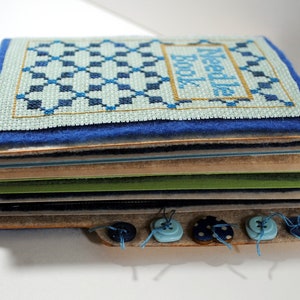 May include: A blue and green needle book with the text "Needle Book" stitched on the front. The book is made of multiple layers of fabric and paper, and has blue buttons attached to the bottom edge.