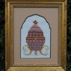 May include: A framed cross-stitch design of a pink and green Easter egg with gold accents. The egg is surrounded by a gold scroll design.