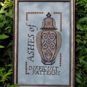 May include: A framed cross-stitch design featuring a blue and gold patterned vase with the text "Ashes of Difficult Patterns".