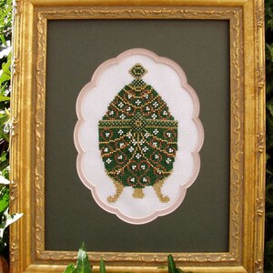 May include: A framed cross-stitch of a green and gold egg with intricate floral designs. The egg is sitting on three gold legs and is surrounded by a white background with a pink border.