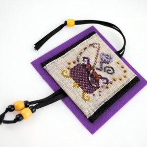 May include: A purple felt pouch with a black ribbon and yellow beads. The pouch has a stitched design of a cauldron with a purple and orange design.