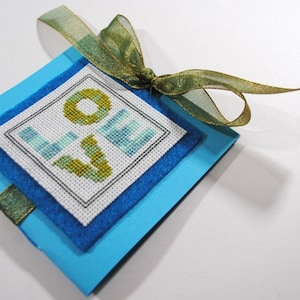 Op de afbeelding: Een blauwe kaart met een groen en blauw gekruist gestikt "LOVE" ontwerp. De kaart is vastgebonden met een goud en groen lint.