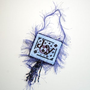 May include: A blue and purple beaded ornament with a cross-stitch design that says "Joy". The ornament is decorated with a fluffy purple fringe.