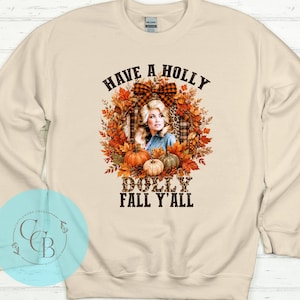 May include: A cream-colored crewneck sweatshirt featuring a graphic print of a woman with blonde hair surrounded by autumn leaves, pumpkins, and a plaid bow. The text reads HAVE A HOLLY DOLLY FALL Y'ALL in black lettering with leopard print accents.