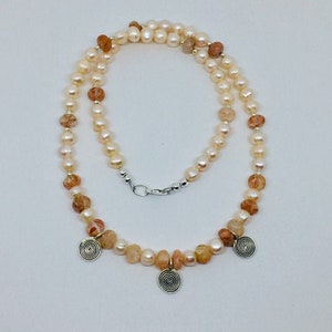 May include: A delicate necklace made with white pearls and orange beads, accented with three silver spiral charms.