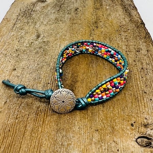 May include: A teal leather bracelet with a colorful beaded design and a silver button closure. The button has a decorative flower design.