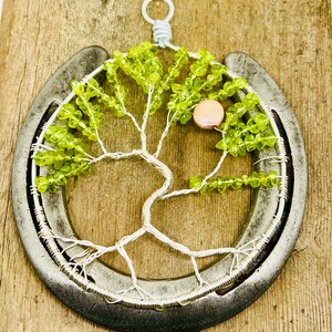 May include: A green glass bead tree of life sculpture is displayed on a horseshoe. The tree is made of wire and has a pink glass bead at the top.