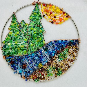 May include: A circular beaded wall hanging with a wire frame. The design features two green trees, a blue river, and a yellow, orange, and red sunset.