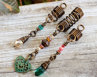 May include: Three bronze-toned hair accessories with spiral accents and decorative beads. One features a green filigree heart charm, another a clear crystal, and the third a green crystal. These accessories are designed for dreadlocks or braids.