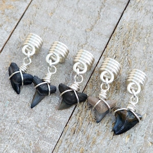 May include: Five shark tooth pendants with silver-colored wire wrapping and coiled tops. The shark teeth vary in color from black to tan and are suspended from silver-colored chain links. The pendants are displayed on a weathered wooden surface.