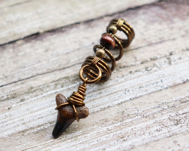 May include: A gold-toned hair coil with a brown and black shark tooth charm. The coil is made of wire and has a spiral design. The charm is attached to the coil with a wire loop.