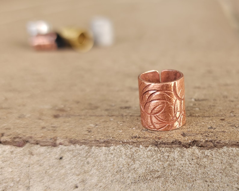 May include: A copper ring with a decorative etched design. The ring is a cylinder shape and has a smooth, polished finish.