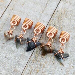 May include: Five shark tooth pendants with copper wire wrapping and copper coil beads. The shark teeth vary in color from light gray to dark gray and black. The pendants are displayed on a weathered wooden surface.
