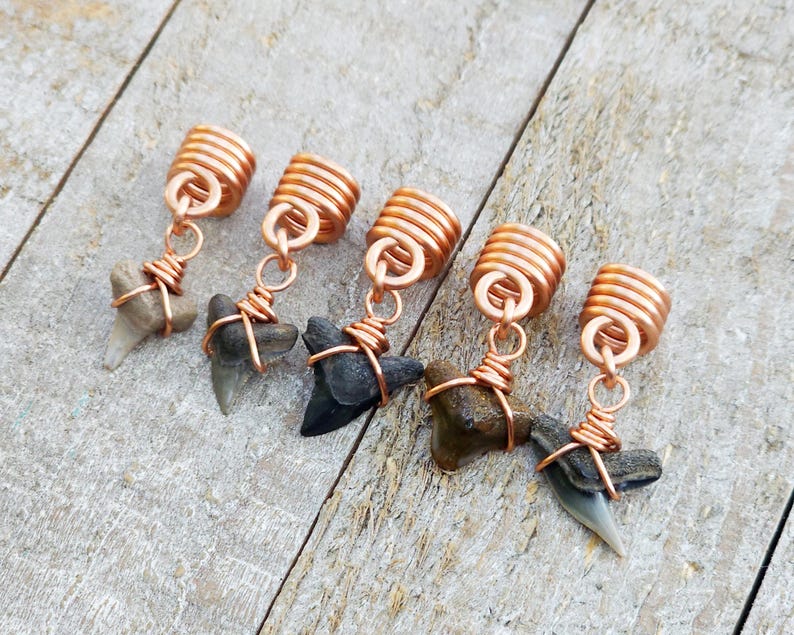 May include: Five shark tooth pendants with copper wire wrapping and copper coil beads. The shark teeth vary in color from light gray to dark gray and black. The pendants are displayed on a weathered wooden surface.