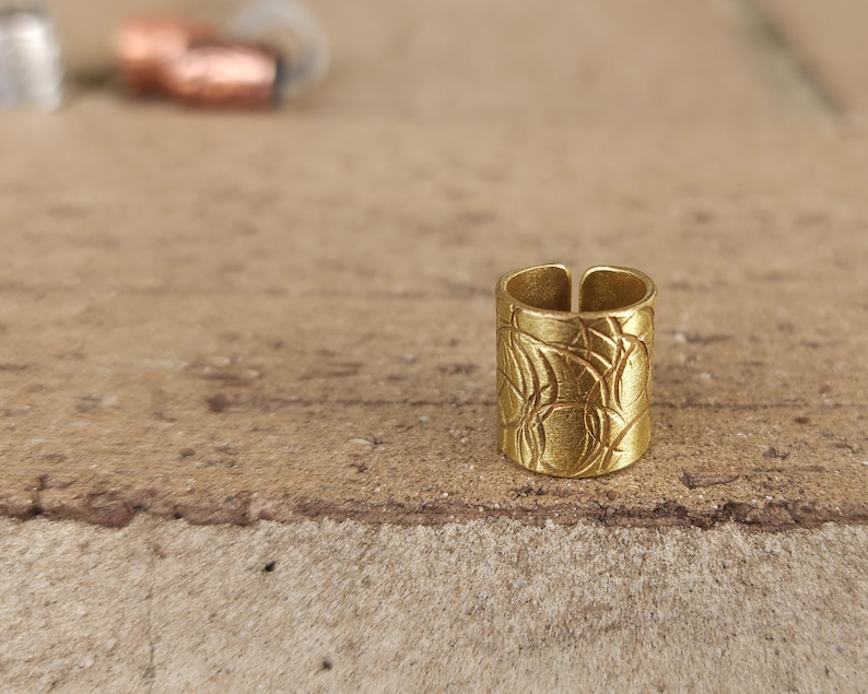 May include: A gold-toned ring with a textured, patterned design. The ring is open on one side and has a slightly curved shape.