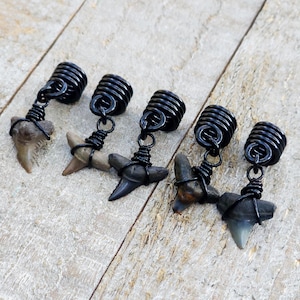 May include: Five shark tooth pendants with black wire wrapping and black coiled tops. The shark teeth vary in color from tan to gray. The pendants are displayed on a light wood surface.