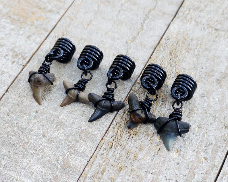 May include: Five shark tooth pendants with black wire wrapping and black coiled tops. The shark teeth vary in color from tan to gray. The pendants are displayed on a light wood surface.
