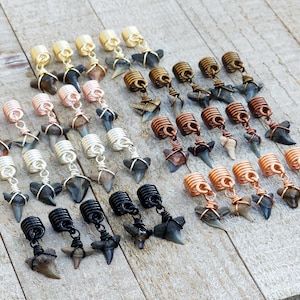 May include: An assortment of handcrafted shark tooth earrings. The earrings feature various wire-wrapped designs in gold, copper, silver, and black, each suspending a natural shark tooth. The earrings are arranged on a light wood surface.