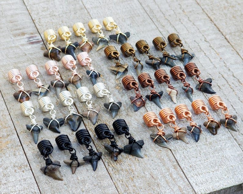 May include: An assortment of handcrafted shark tooth earrings. The earrings feature various wire-wrapped designs in gold, copper, silver, and black, each suspending a natural shark tooth. The earrings are arranged on a light wood surface.