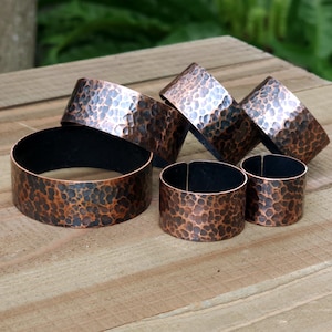 May include: A collection of copper napkin rings with a hammered texture. The rings vary in size and are arranged on a wooden surface. The copper has a rich, warm color with dark accents, and some rings have a black interior.