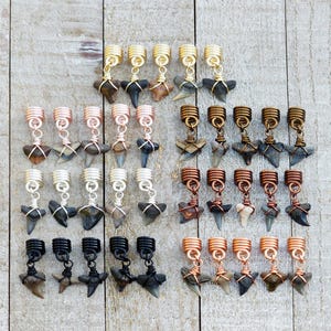 May include: Collection of hair jewelry featuring shark teeth pendants in various metallic finishes, including gold, copper, silver, and black. The pendants are attached to coiled metal hair beads, offering a unique accessory.