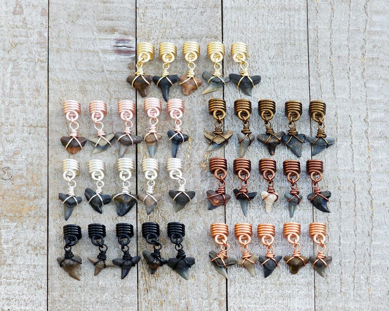 May include: Collection of hair jewelry featuring shark teeth pendants in various metallic finishes, including gold, copper, silver, and black. The pendants are attached to coiled metal hair beads, offering a unique accessory.