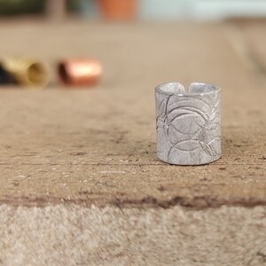 May include: A silver ring with a textured design. The ring is made of metal and has a circular shape.