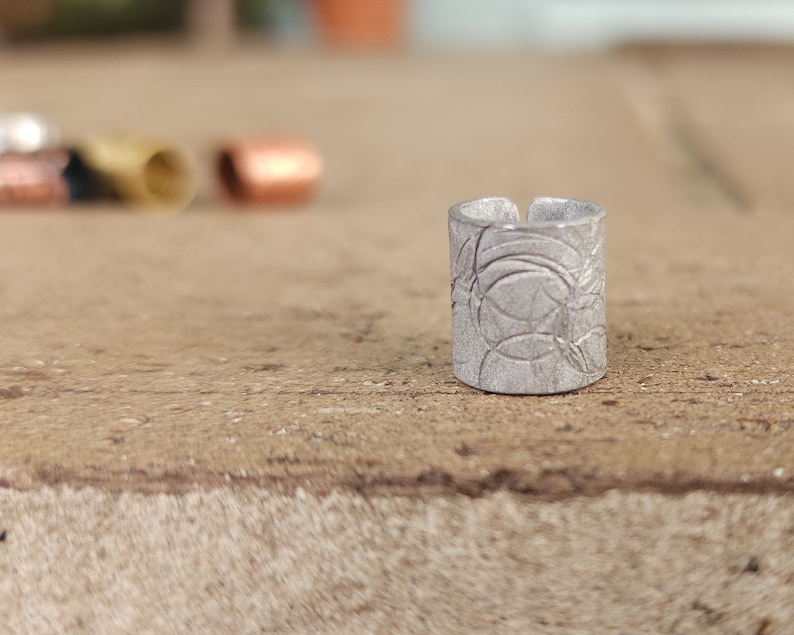 May include: A silver ring with a textured design. The ring is made of metal and has a circular shape.