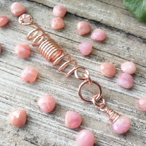 May include: A rose gold hair coil with a pink opal gemstone at the top and bottom. The coil is made of wire and has a spiral shape. The gemstones are faceted and have a shimmery finish.