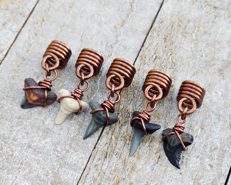 May include: Five handcrafted hair accessories featuring fossilized shark teeth. Each tooth is unique in color, ranging from brown to gray, and is wrapped with copper wire. The hair accessories have copper coil accents.