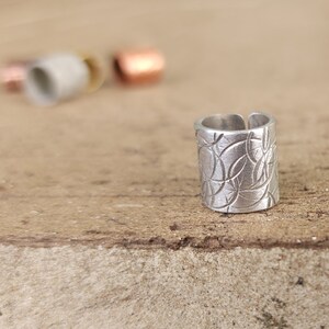 May include: A silver ring with a textured design. The ring is made of metal and has a cylindrical shape.
