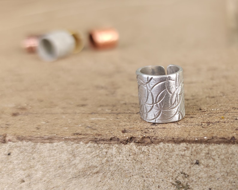 May include: A silver ring with a textured design. The ring is made of metal and has a cylindrical shape.