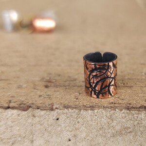 May include: A copper ring with a black, abstract design. The ring is made of metal and has a textured surface.