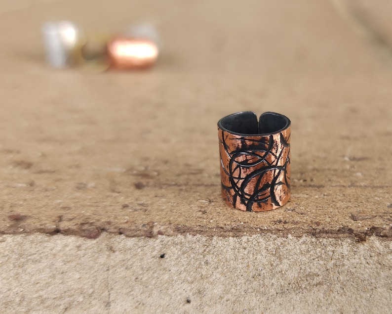 May include: A copper ring with a black, abstract design. The ring is made of metal and has a textured surface.