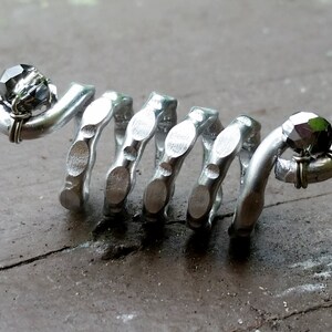 May include: A silver metal ring with a spiral design and two black beads. The ring is made of recycled materials and is a unique piece of jewelry.