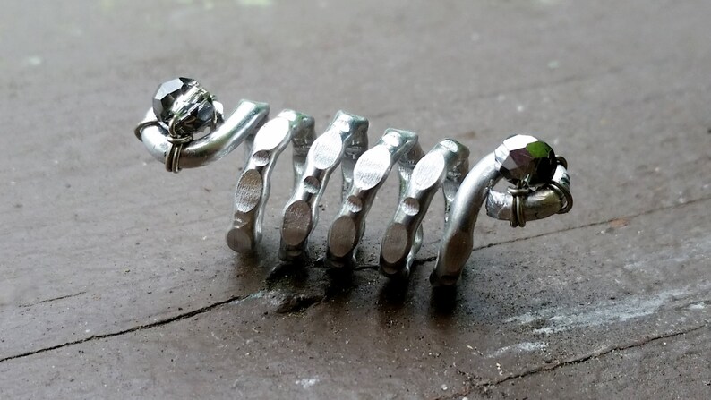 May include: A silver metal ring with a spiral design and two black beads. The ring is made of recycled materials and is a unique piece of jewelry.