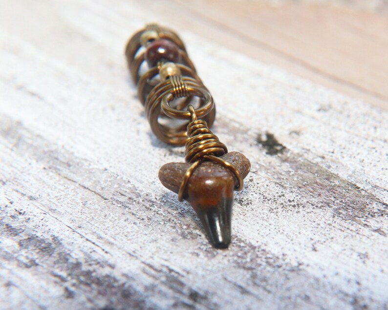 May include: A brown and gold shark tooth pendant with wire wrapped beads. The pendant is on a white wooden surface.