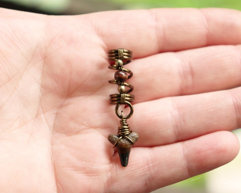 May include: A gold-toned hair coil with a brown and white shark tooth charm. The coil is made of wire and has a spiral design.