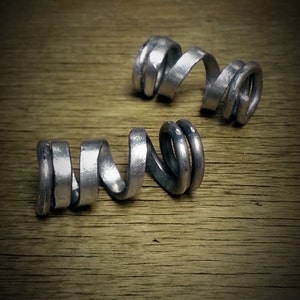 Puede incluir: Dos anillos de metal plateado, cada uno enrollado en forma de espiral. Los anillos están sobre una superficie de madera.