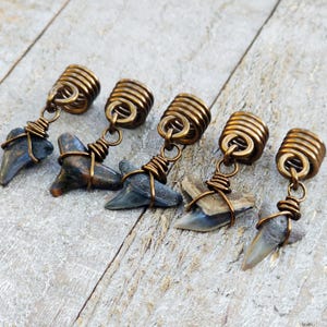 May include: Five handcrafted shark tooth pendants. Each pendant features a natural shark tooth wrapped in copper wire, suspended from a coiled brass bead. The shark teeth vary in color, with shades of gray, brown, and cream.