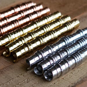 Puede incluir: Filas de objetos metálicos en forma de espiral en tonos cobre, dorado y plateado. Los objetos están dispuestos en líneas paralelas, con una superficie de madera en el fondo. El diseño en espiral es consistente en todos los objetos.