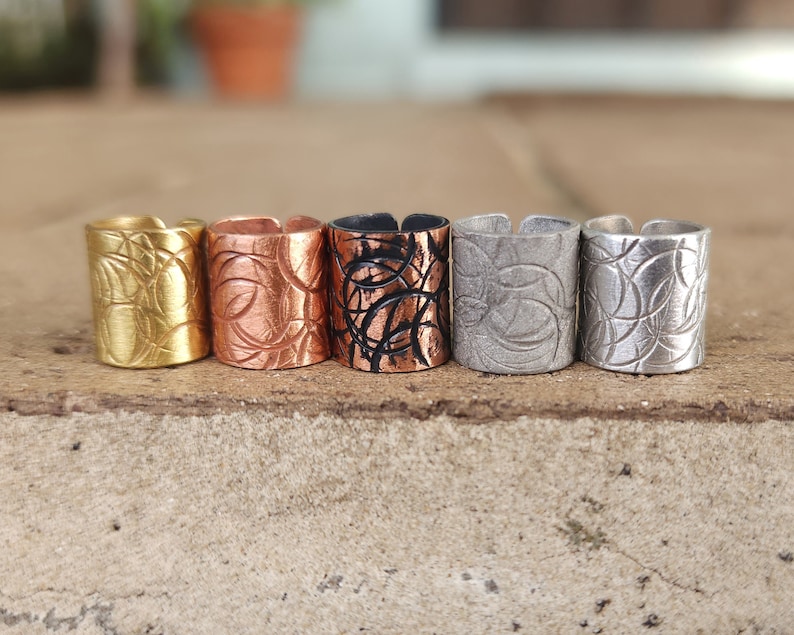 May include: Five metal rings with a textured design. The rings are in different colors: gold, copper, black, silver, and white.